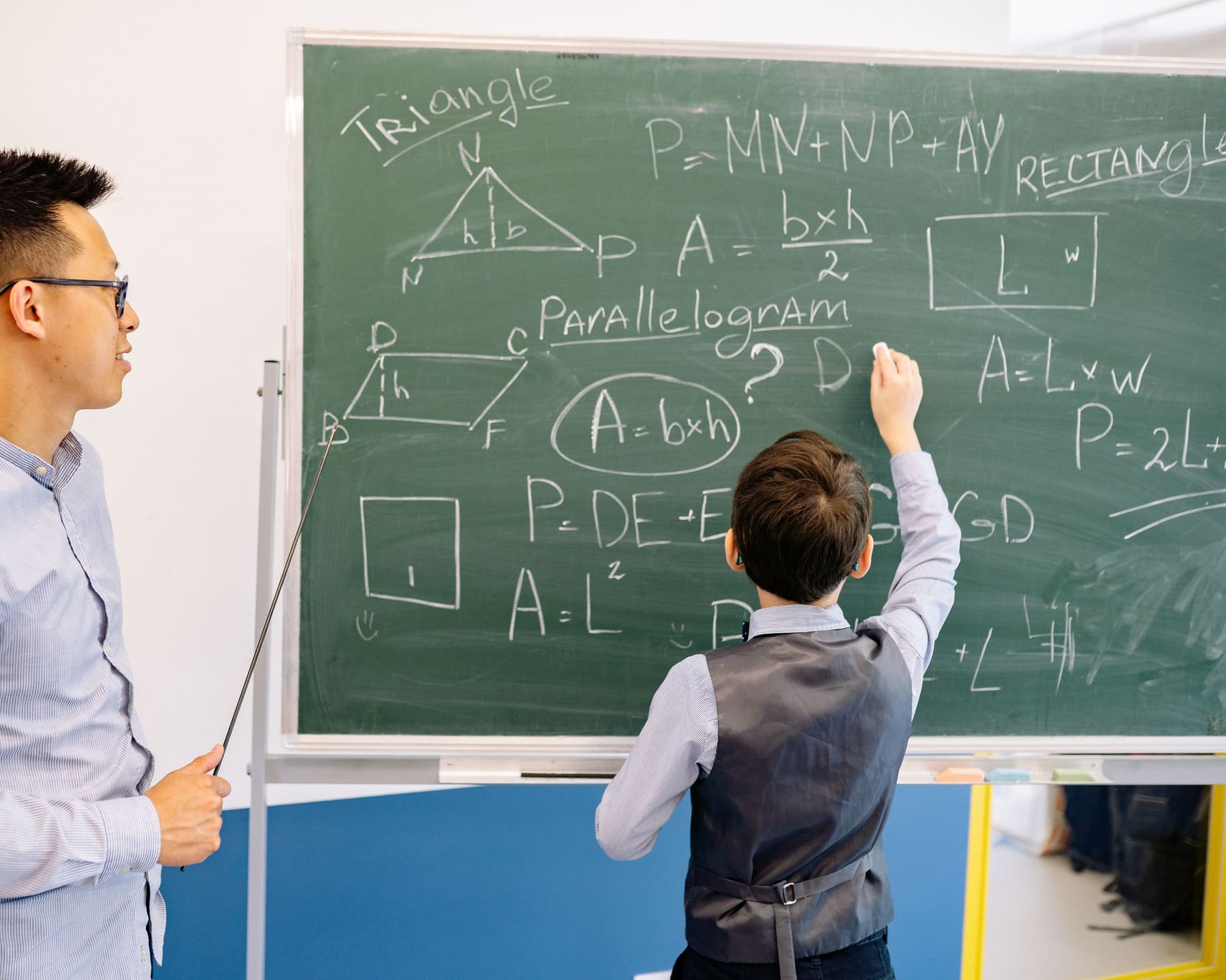 A student working through a geometry problem on a chalkboard while a teacher looks on